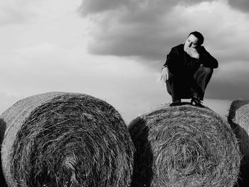 Full length of man crouching on hay bale against cloudy sky