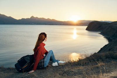 Man sitting on rock by lake against sky during sunset