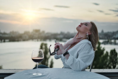 Side view of young woman holding drink against sky at sunset