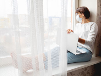 Woman in medical mask remote works from home. she sits with laptop on knees. quarantine.