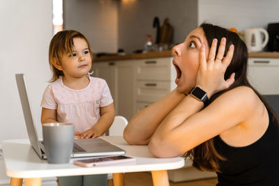 Toddler looking at shocked mother with laptop at home