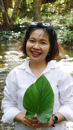 Portrait of cheerful woman holding leaf while standing at riverbank in forest