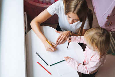 Little girl toddler with her mother drawing with colored pencils on table in children's room at home