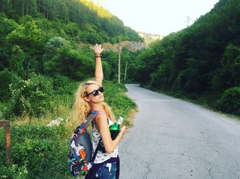 Young woman carrying backpack and beer bottle standing on road against mountains