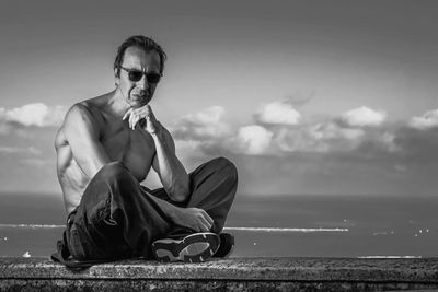 Shirtless mature man sitting against sea and sky