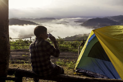 Rear view of young man having drink while sitting at campsite against sky