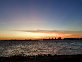 Silhouette cranes at harbor against sky during sunset