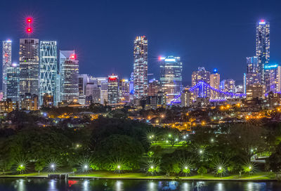 Illuminated modern buildings by trees against sky in city at night