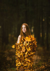 Thoughtful young woman covered with leaves standing at forest during autumn