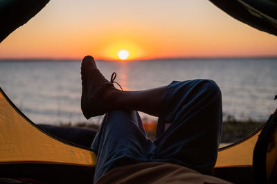 Low section of man relaxing in tent by sea against sky during sunset