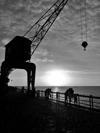 Silhouette people on pier crane over sea against sky during sunset