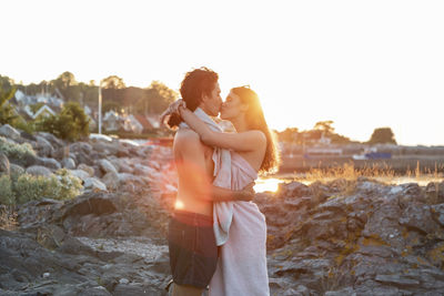 Young couple kissing while standing on rock at seashore during sunset