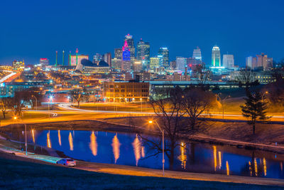 Illuminated buildings by river against clear blue sky at night