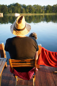 Back of man with straw hat and dog sitting on a wooden chair looking at calm lake at sunset