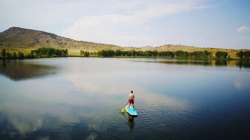 Rear view of shirtless young man paddleboarding on lake against sky