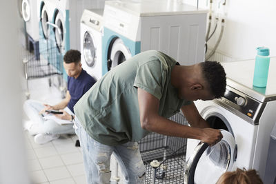 Man using washing machine while university student studying at laundromat