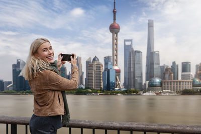 Smiling woman photographing cityscape by river against sky