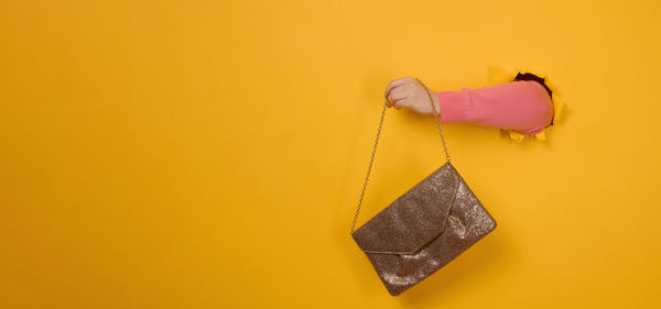 Female hand is holding a small golden clutch bag with a metal chain on a yellow background. part of 