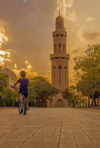 Man riding bicycle by building against sky during sunset