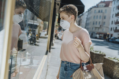 Woman wearing protective face mask doing window shopping while standing on street in city