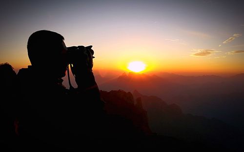 Side view of silhouette man photographing with camera on cliff at sunset