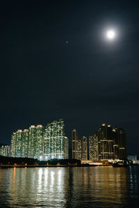 Illuminated modern buildings by sea against sky at night