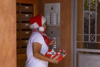 Senior woman wearing mask holding gift standing by door