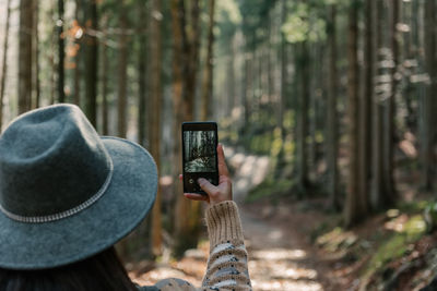 Rear view of woman holding mobile phone, taking photos of moody forest in autumn