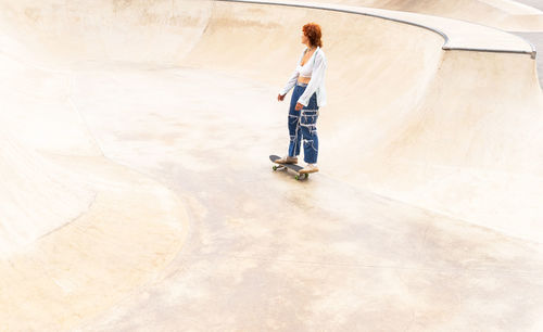 From above full body side view of active female in casual wear riding skateboard in skate park with ramp on summer day