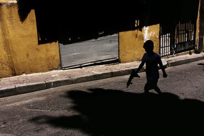 Silhouette boy with toy gun walking on road during sunny day