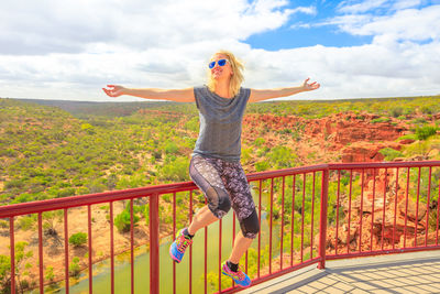 Full length of woman with arms outstretched sitting against landscape on railing at kalbarri national park