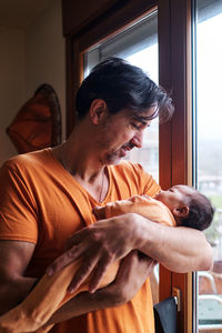 Bearded mature hispanic dad gently holding cute little baby while standing near glass door at home