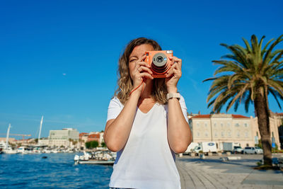 Portrait of young woman photographing sea against clear sky