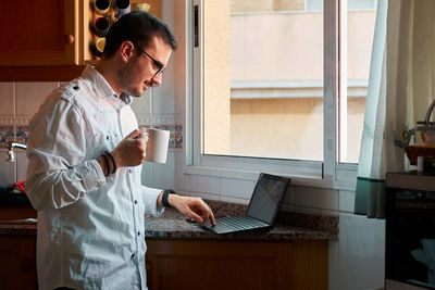 Young man looks at his laptop while drinking coffee in his kitchen