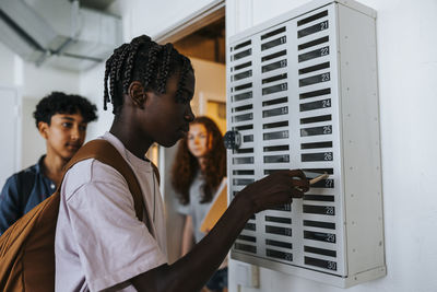 Teenage student with braided hair keeping smart phone in locker at junior high school