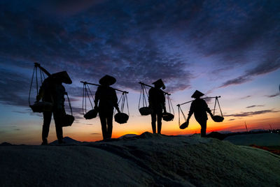 Silhouette people with baskets while standing on land against sky during sunset