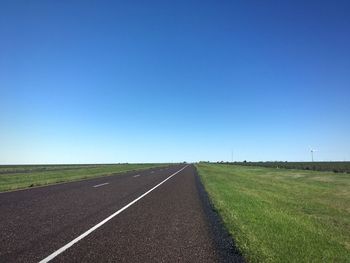 Empty road amidst grassy landscape against clear blue sky