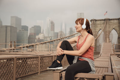 Woman listening to music while sitting on bridge against sky