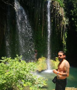 Full length of shirtless man standing against waterfall