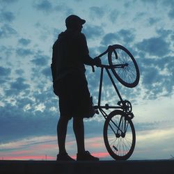 Low angle view of silhouette man standing on bicycle against sky