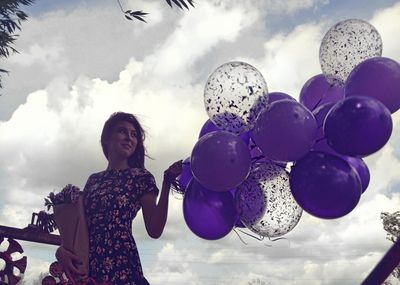 Low angle view of smiling woman holding blue balloons against cloudy sky