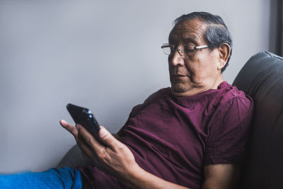 Senior ethnic male in glasses relaxing on couch in living room and reading messages on smartphone