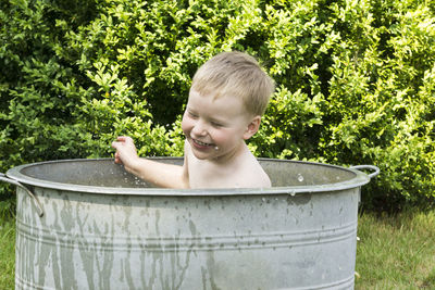 Cheerful shirtless boy taking bath while sitting in container at yard