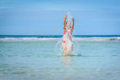 Happy mid adult woman splashing water in sea against sky