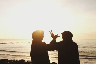 Silhouette friends gesturing peace sign while standing on beach against clear sky during sunset