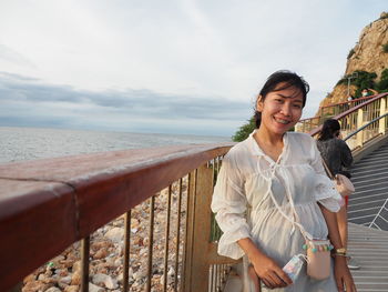Smiling young woman standing by railing against sea