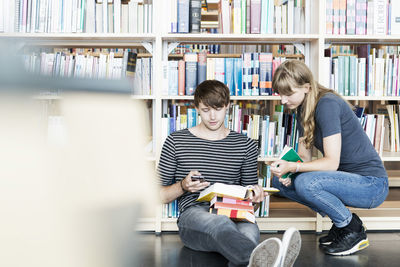 Full length of young students with books using mobile phone in college library