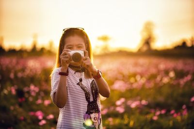 Woman photographing with camera while standing on field during sunset