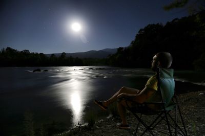 Man sitting on lakeshore against sky at night