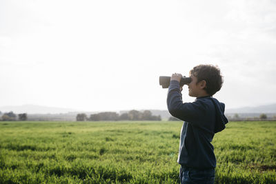 Boy looking through binoculars at agricultural field during sunny day